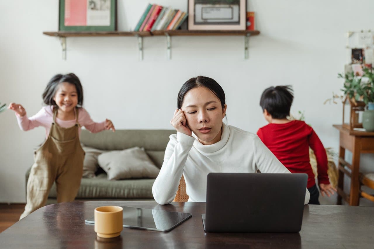 A woman at a laptop at home while young children play behind her, representing women trying to work and manage childcare
