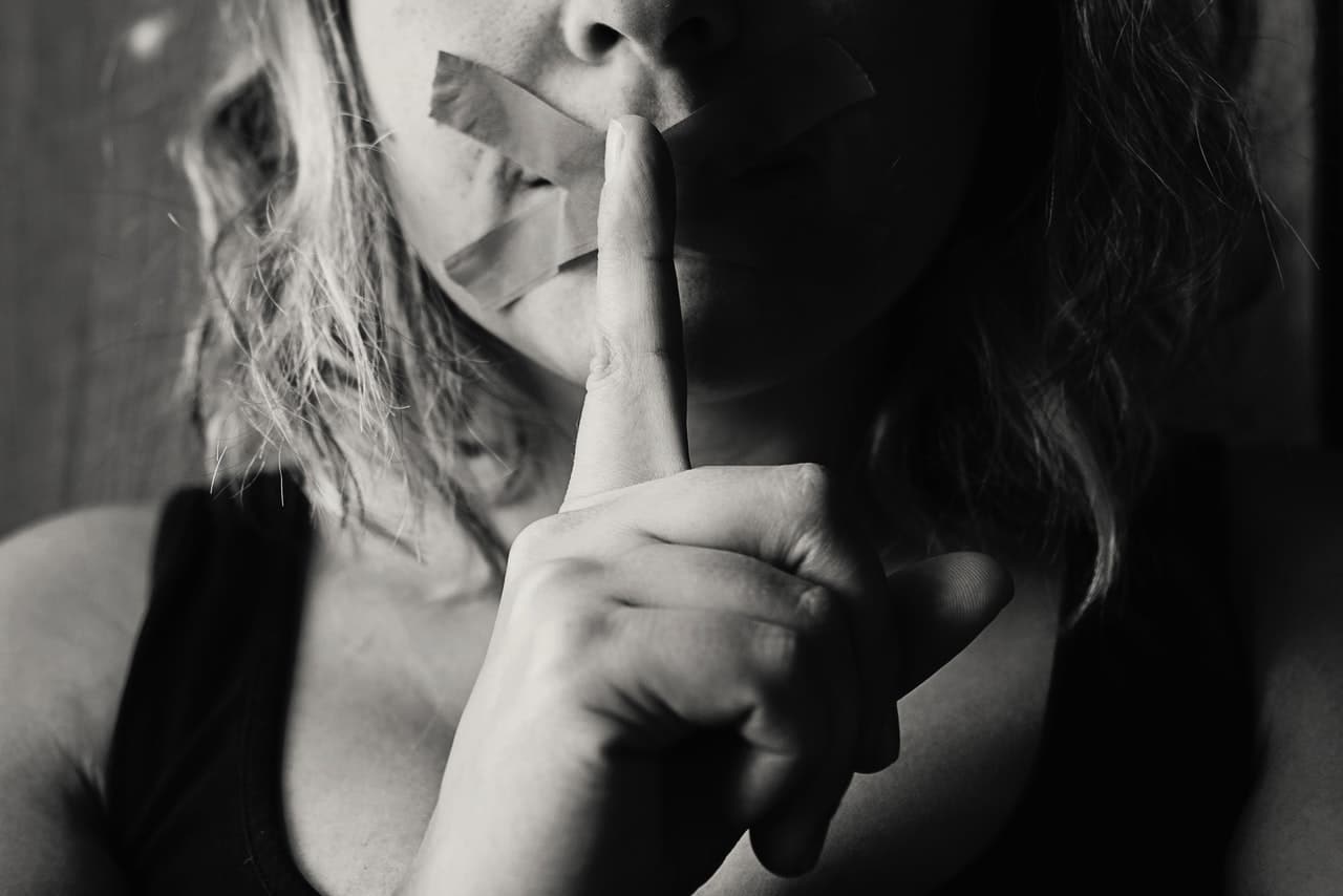 A close up of a woman with tape over her mouth holding a finger to her lips representing staying silent after abuse