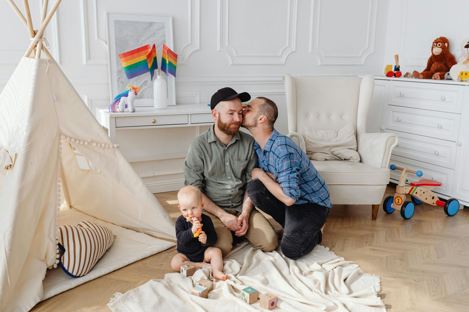 A man kissing his male partner on the cheek with their baby in a child's bedroom, representing surrogacy and LGBTQIA2S+ parents