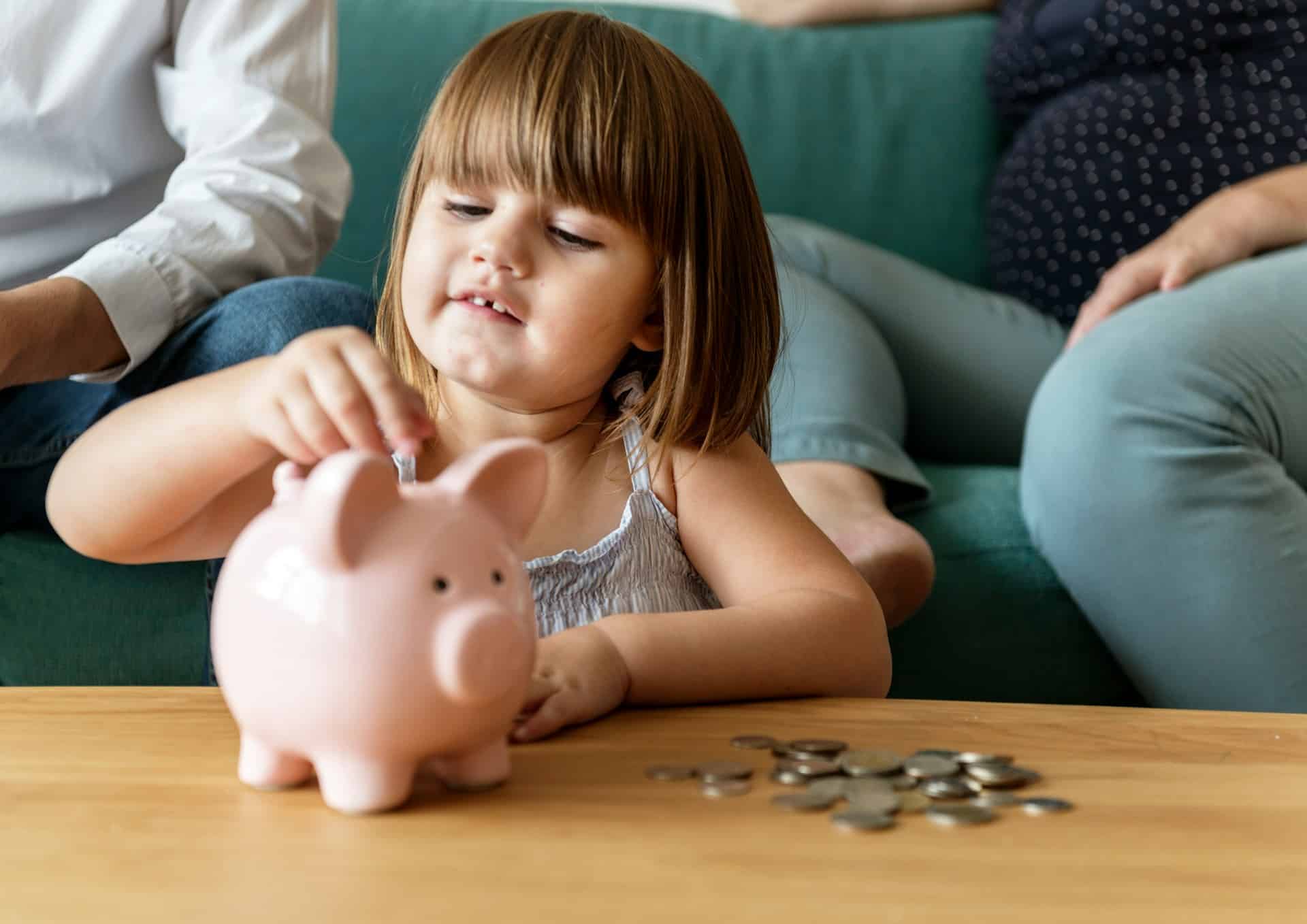 A toddler girl placing coins into a piggy bank while her parents watch from the couch, representing property division, spousal support and child support in Alberta